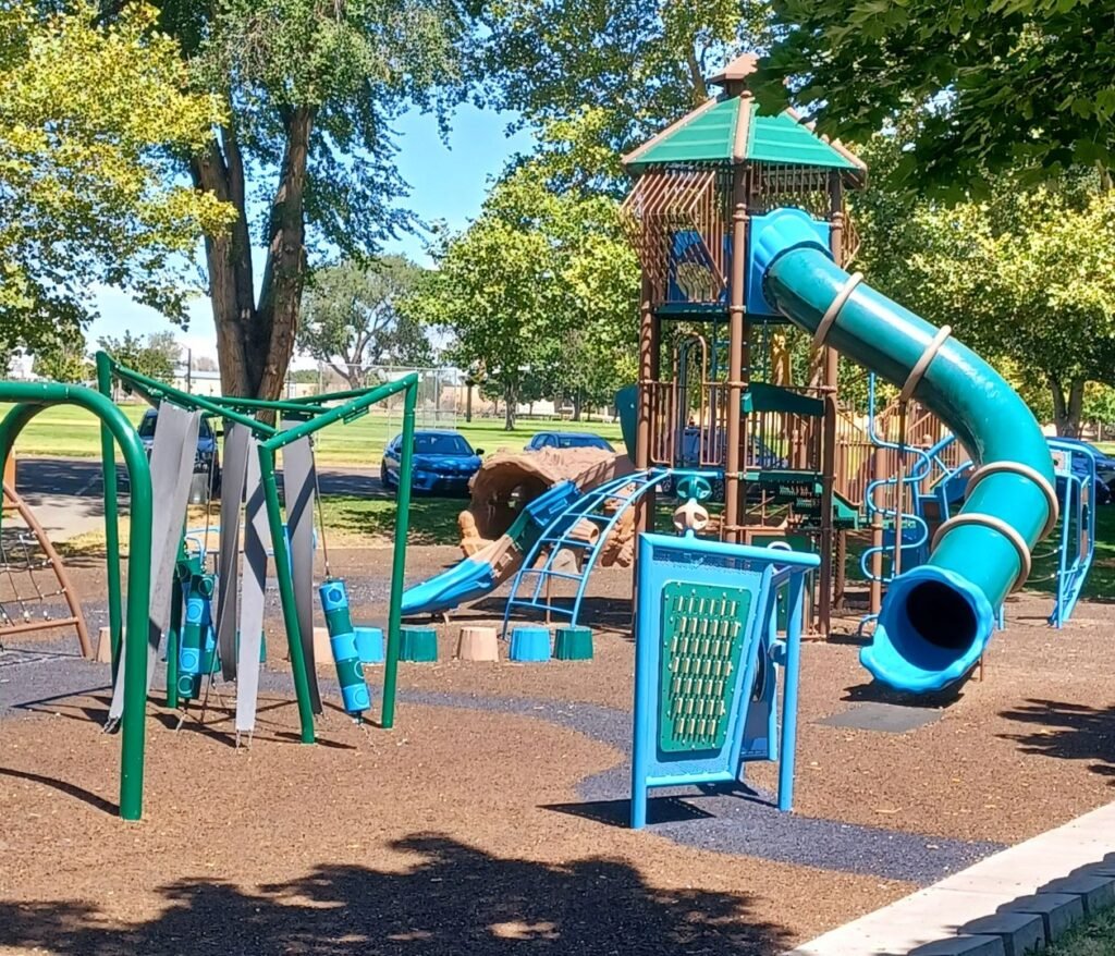 Lakeview Park playground in Nampa, Idaho, with a large blue tube slide, climbing structures, swings, and shaded trees.